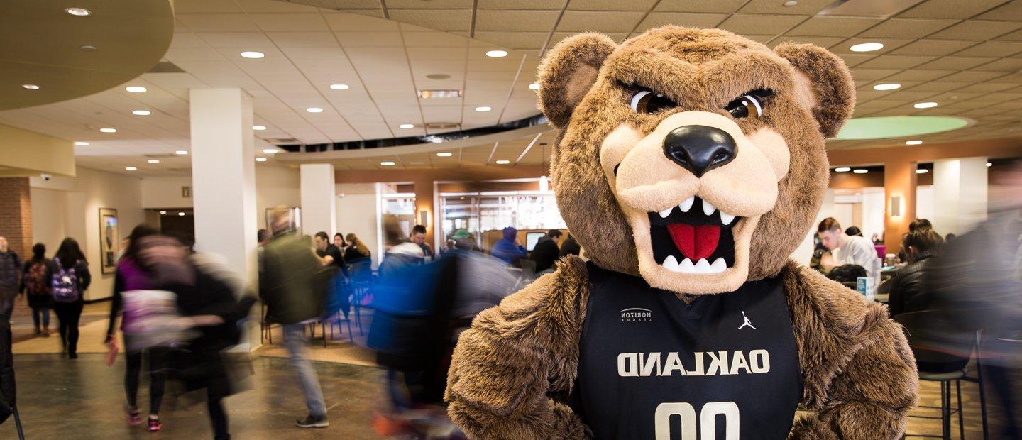 The Grizz bear wearing a jersey that says Oakland on it, with people walking through the room behind him